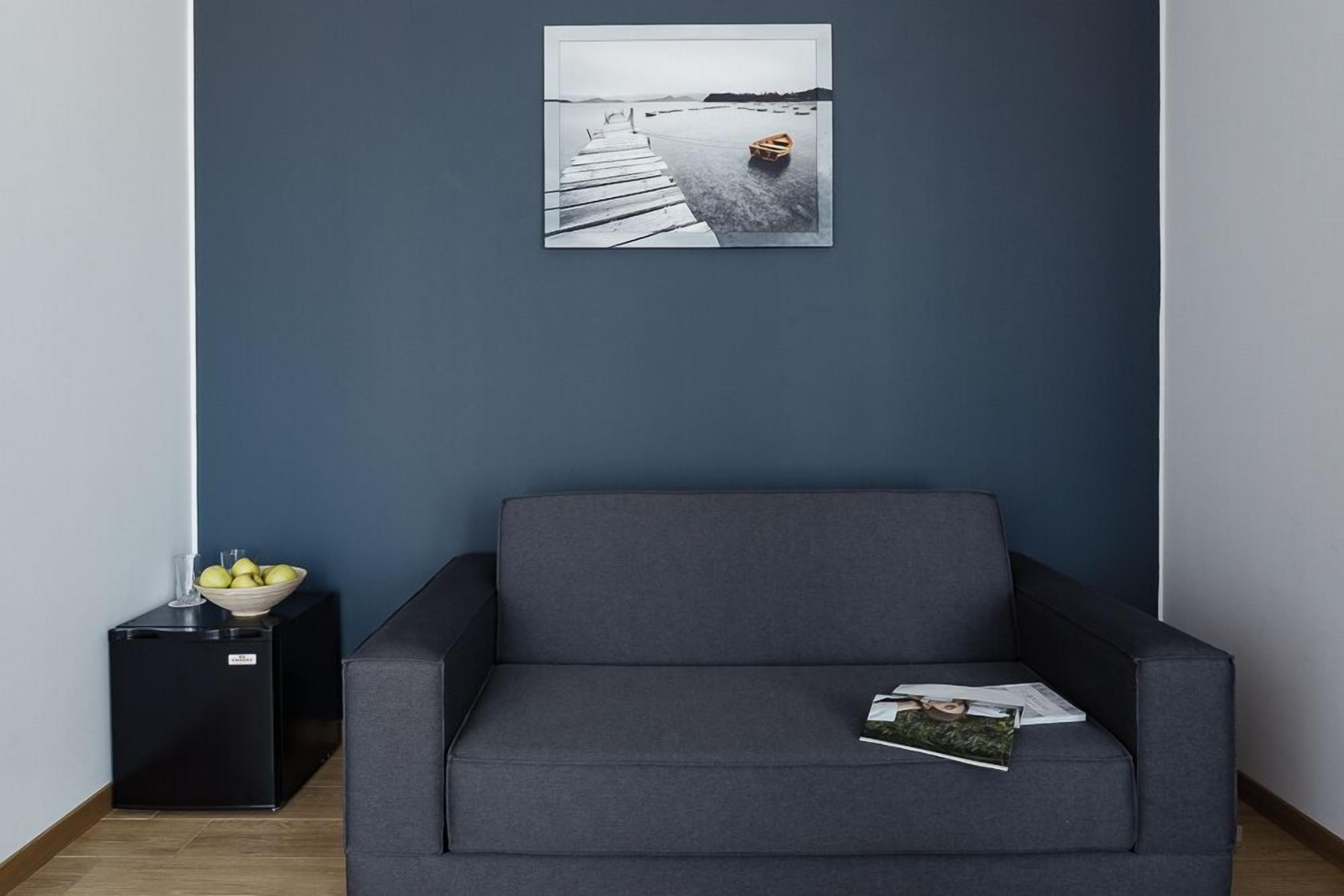 A dark blue room with a grey sofa, a small black side table with a bowl of fruit, and a framed picture of a boat on water.