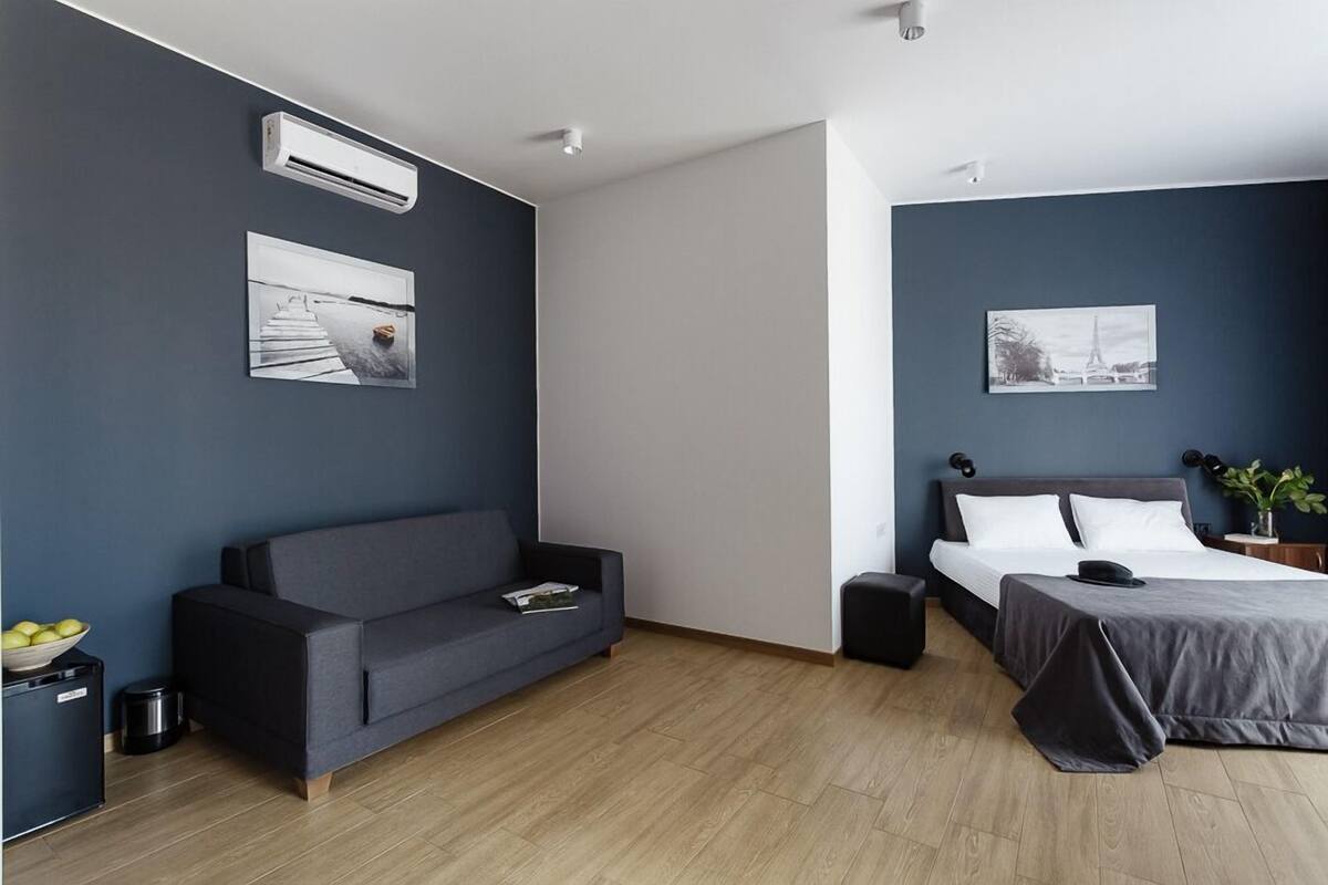 Une chambre d’hôtel moderne avec un canapé gris, un lit recouvert d’une courtepointe grise, une petite table avec une plante et deux unités de climatisation fixées au mur.