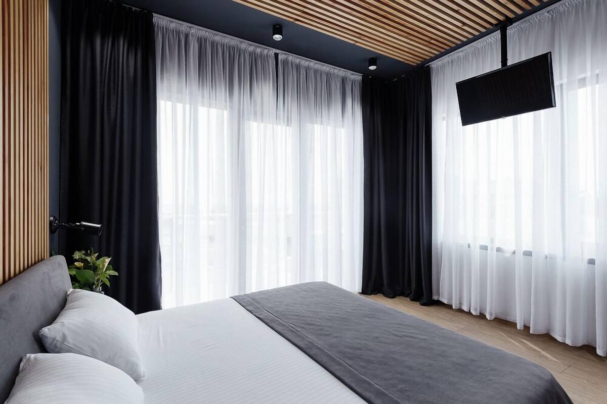 Une chambre d’hôtel moderne dotée d’un grand lit, de rideaux occultants, d’un téléviseur fixé au mur et d’un plafond en bois.