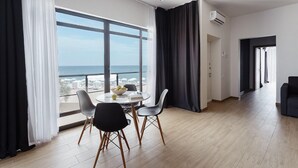 A modern living room with a large window, a dining area with a round table and chairs, and a view of the sea.