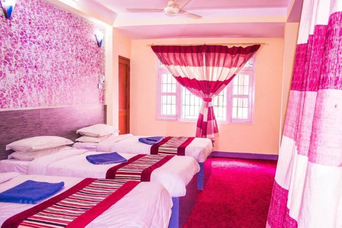 Een hotelkamer met drie bedden, een plafondventilator, een raam met een rood-wit gordijn en een roze vloerkleed.