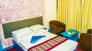 Una habitación de hotel con una cama, un sillón marrón, una mesita con un libro y una ventana con cortinas amarillas.