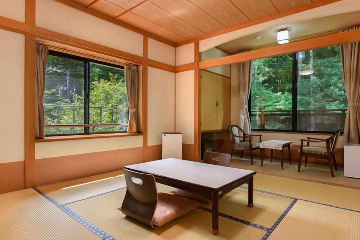Una habitación japonesa tradicional con suelo de tatami, una mesa baja y una silla. Hay dos ventanas con cortinas, una pequeña mesa con sillas y vistas a la vegetación exterior.