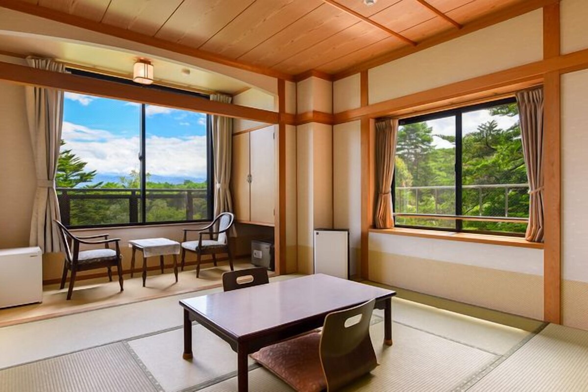 Una habitación tradicional japonesa con suelo de tatami, una mesa baja y sillas. Amplias ventanas ofrecen vistas al follaje.