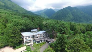 Un hotel moderno con techo plano, varios pisos y un estacionamiento, rodeado de un bosque denso y montañas.