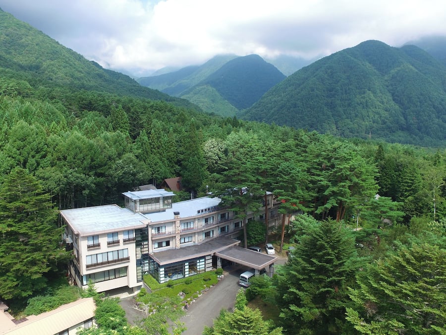 A modern hotel with a flat roof, multiple floors, and a parking area, surrounded by dense forest and mountains.