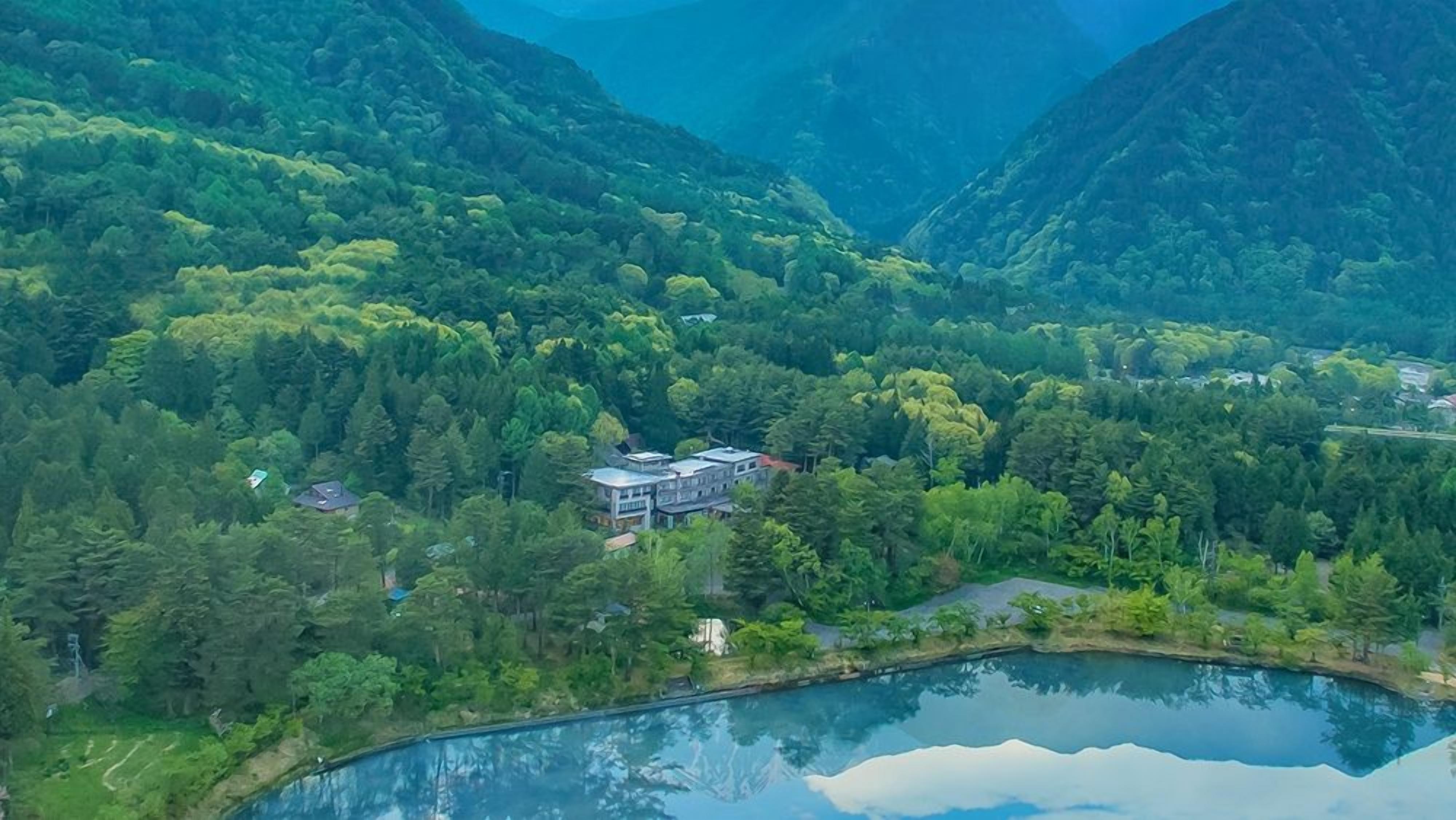 A mountainous landscape with a lake, a house surrounded by dense forest, and a clear sky.