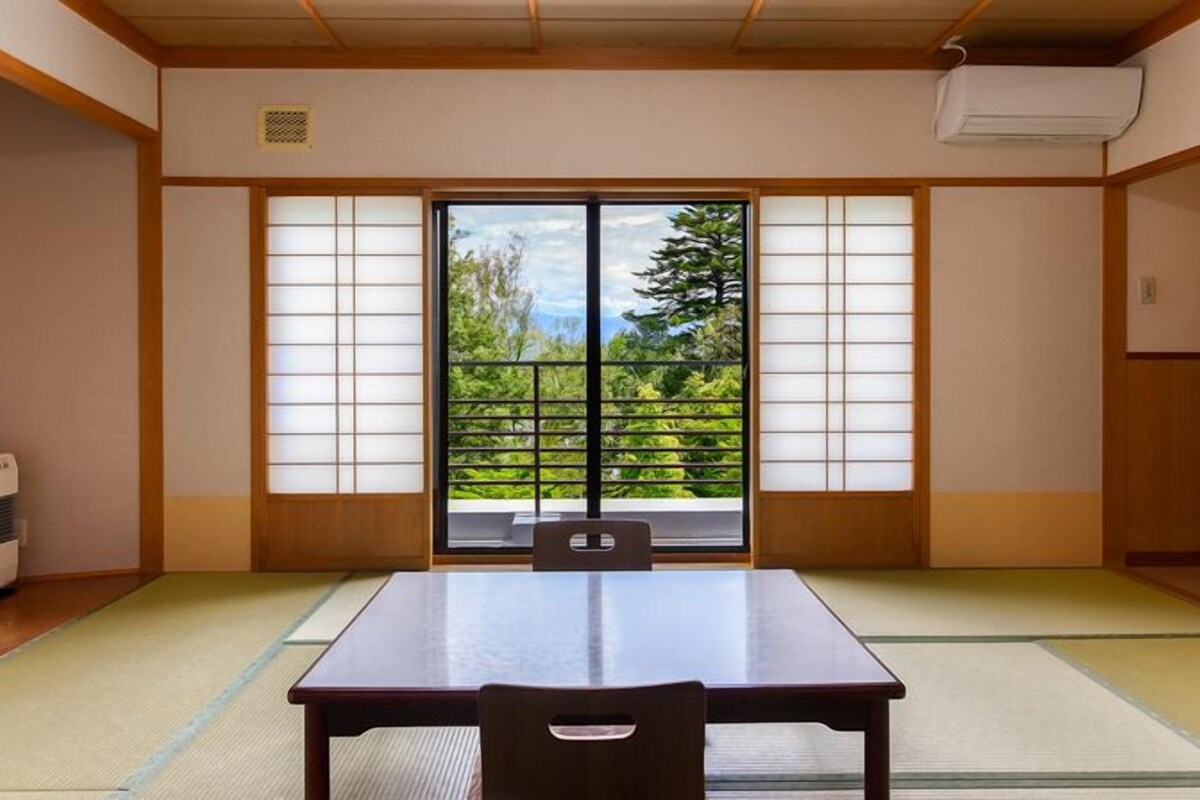 Una habitación japonesa tradicional con suelo de tatami, un fusuma que ofrece vistas a la vegetación y una mesa con una silla.