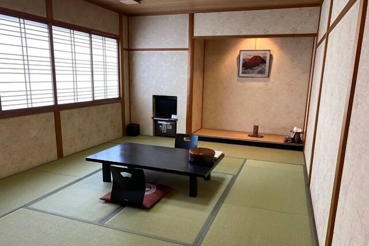 Ein Raum mit Tatami-Matten, einem niedrigen Tisch, einem Fernseher und einem gerahmten Bild an der Wand.