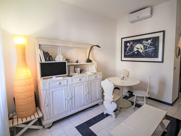 A room with a white cabinet, a flat-screen TV, a lamp, a framed abstract painting, and a round table with chairs.