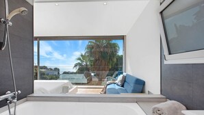 A modern bathroom with a large bathtub, a shower, and a glass-enclosed seating area with a view of palm trees.