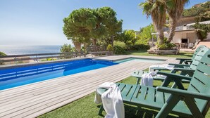 A swimming pool with a wooden deck, green lounge chairs, and a view of the sea.