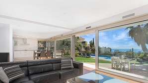 A modern living room with a black leather sofa, a glass coffee table, and a view of a pool and garden through large sliding glass doors.
