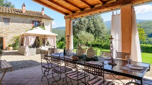 A covered outdoor dining area with a table set for two, surrounded by stone buildings and a scenic view of hills. - Villa Montanina - Relax in Tuscany (Castel Focognano)