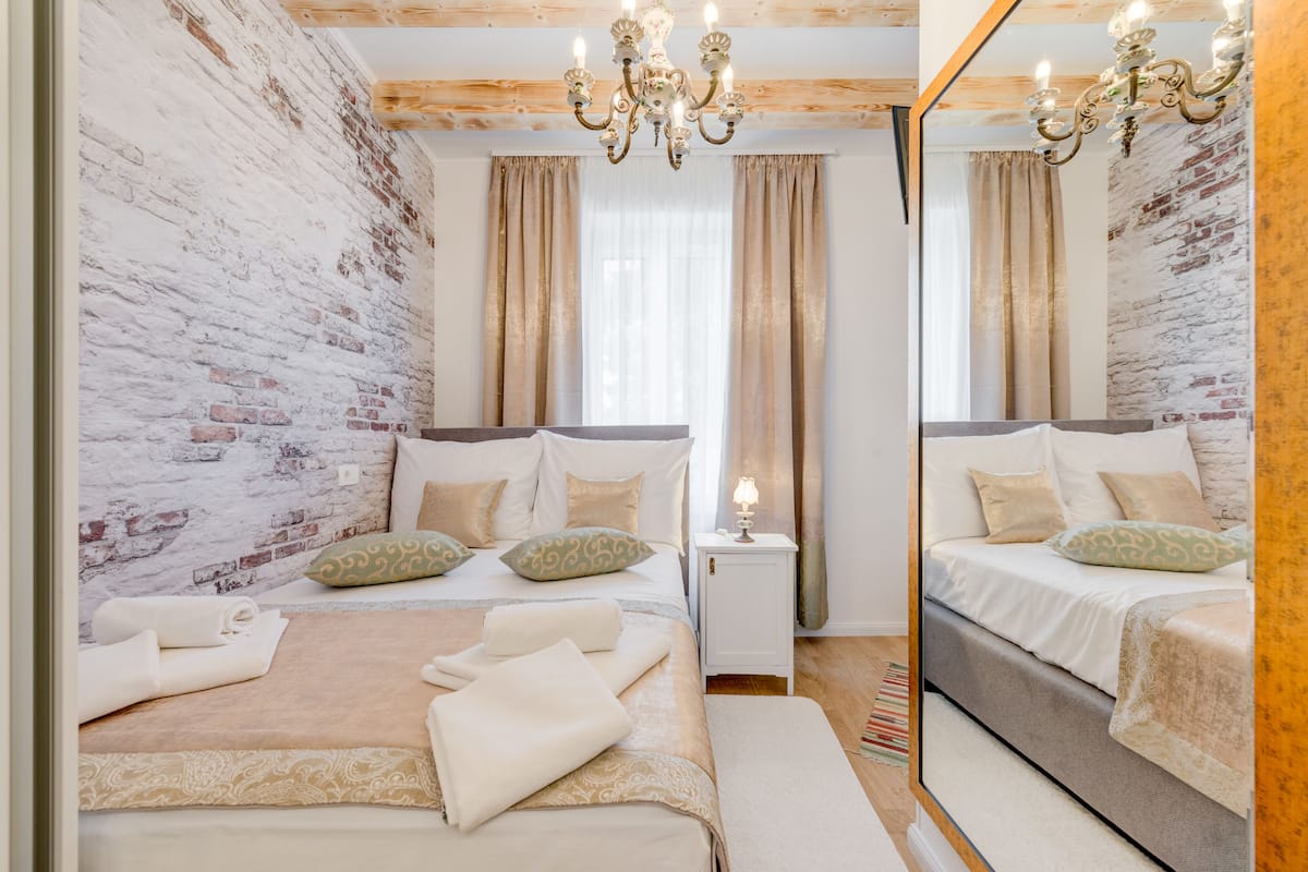 A bedroom with a brick wall, a chandelier, a bed with pillows, a nightstand, and a mirror.