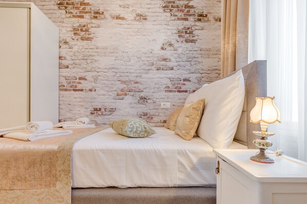 Un dormitorio con pared de ladrillo, cama con ropa de cama blanca, mesita de noche con lámpara y ventana con cortinas.