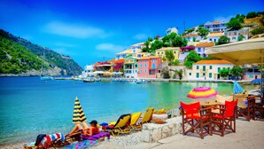 A coastal scene with colorful buildings, a beach with lounge chairs and umbrellas, and boats in the water. - Villa Anthia (Kefalonia)