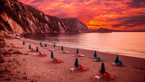 A beach with rows of sun loungers and umbrellas, a rocky cliff, and a sunset over the water. - Villa Anthia (Kefalonia)