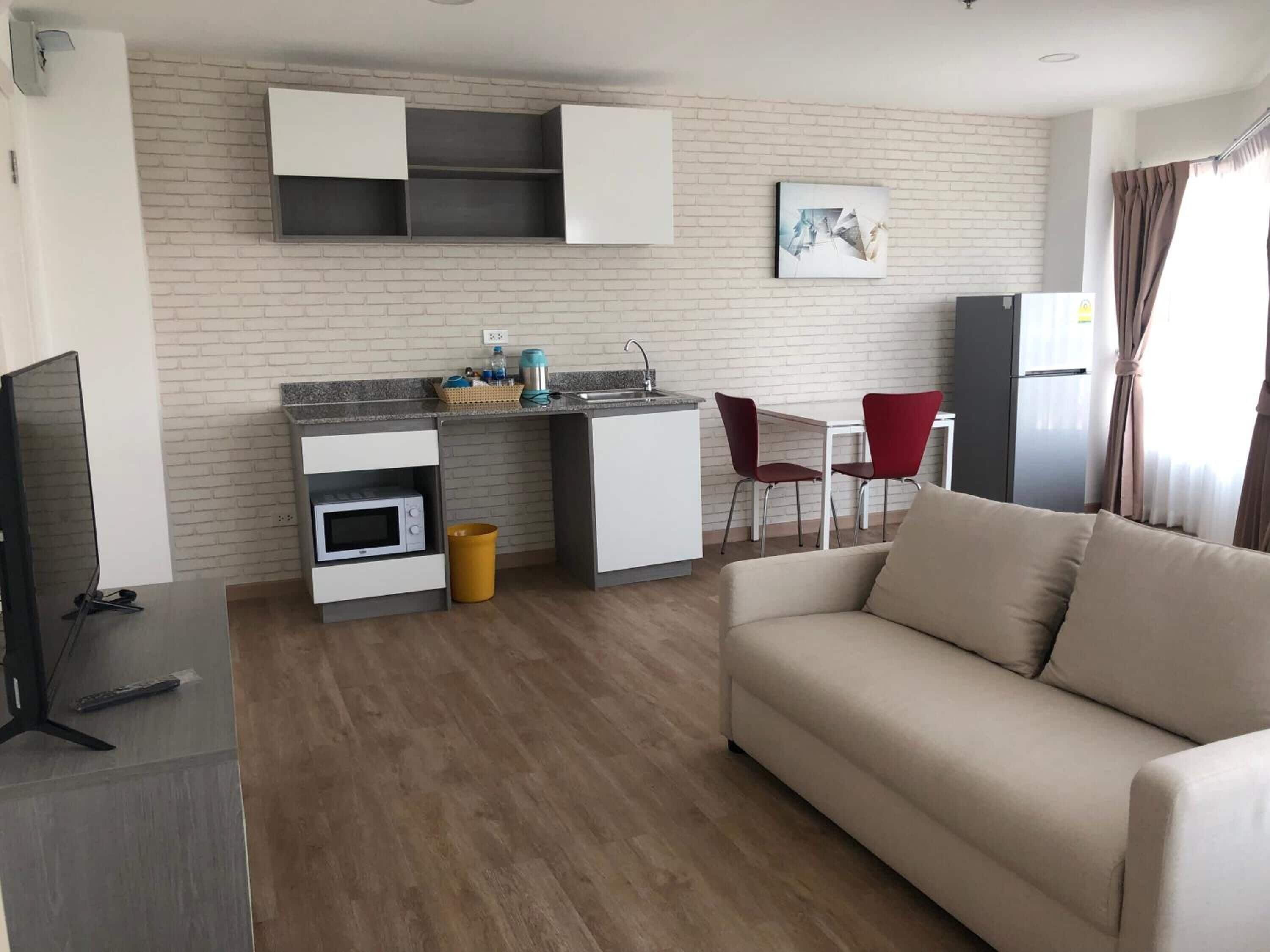 A modern living room with a sofa, a television, a kitchen area with cabinets and a microwave, and a dining area with chairs.