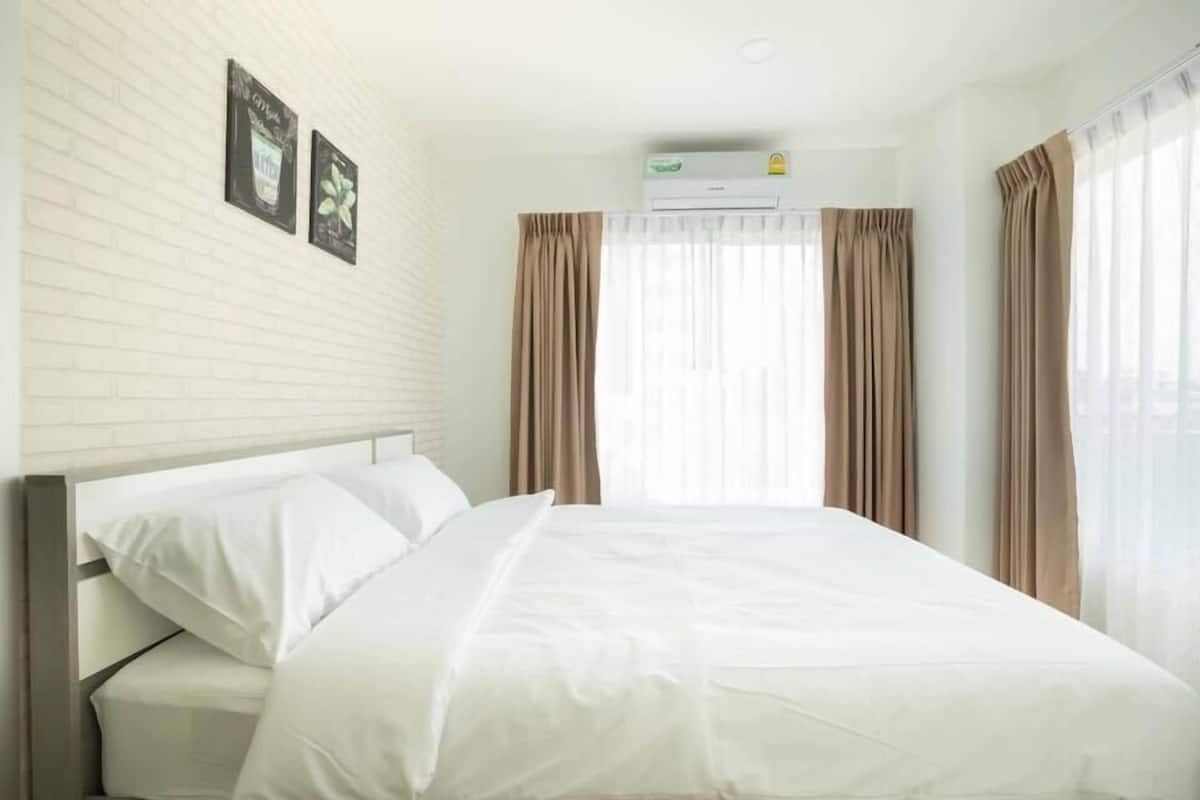A hotel room with a bed, white walls, a brick accent wall, an air conditioning unit, and curtains.