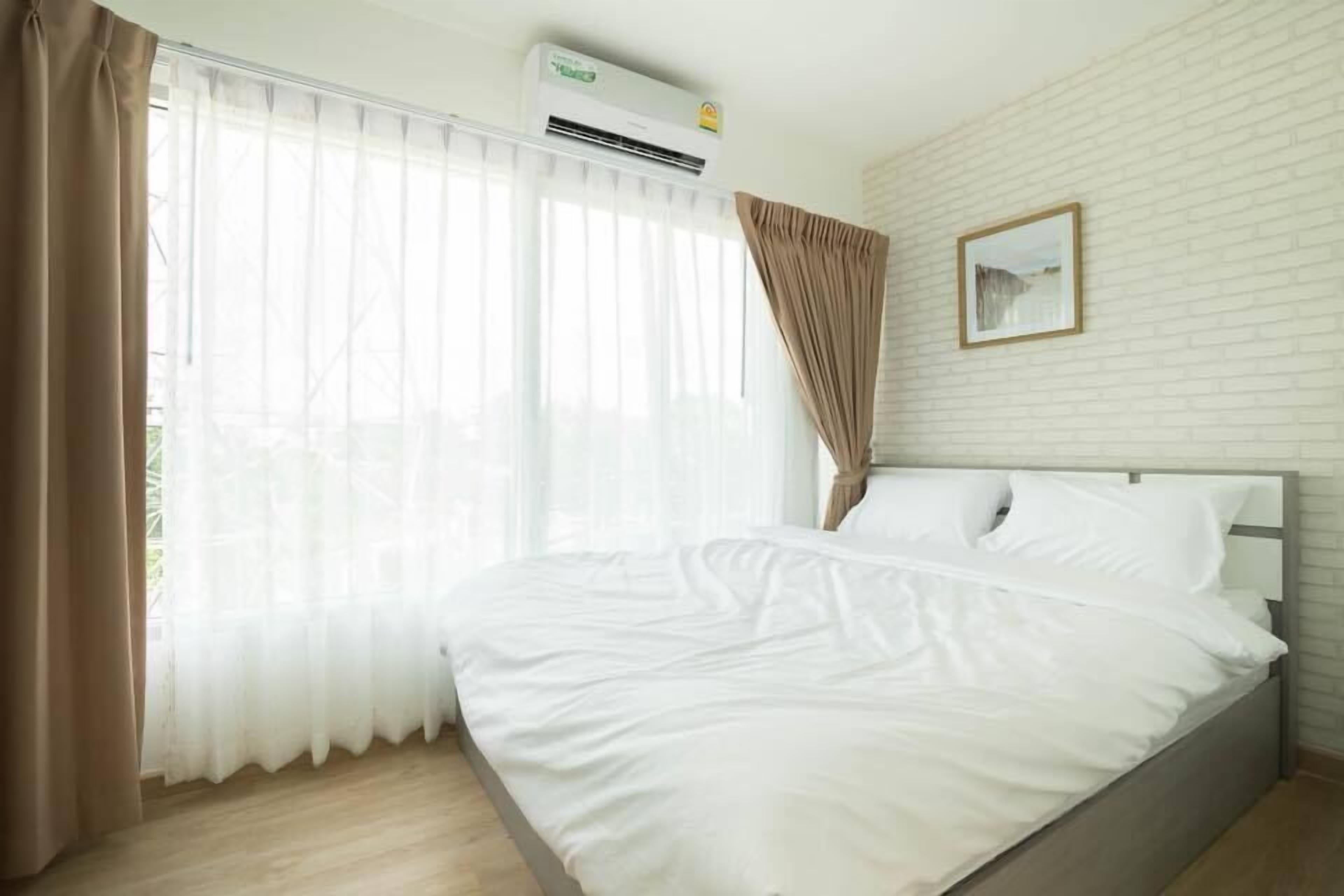 A neatly made bed in a hotel room with a large window, an air conditioning unit, and a picture on the wall.