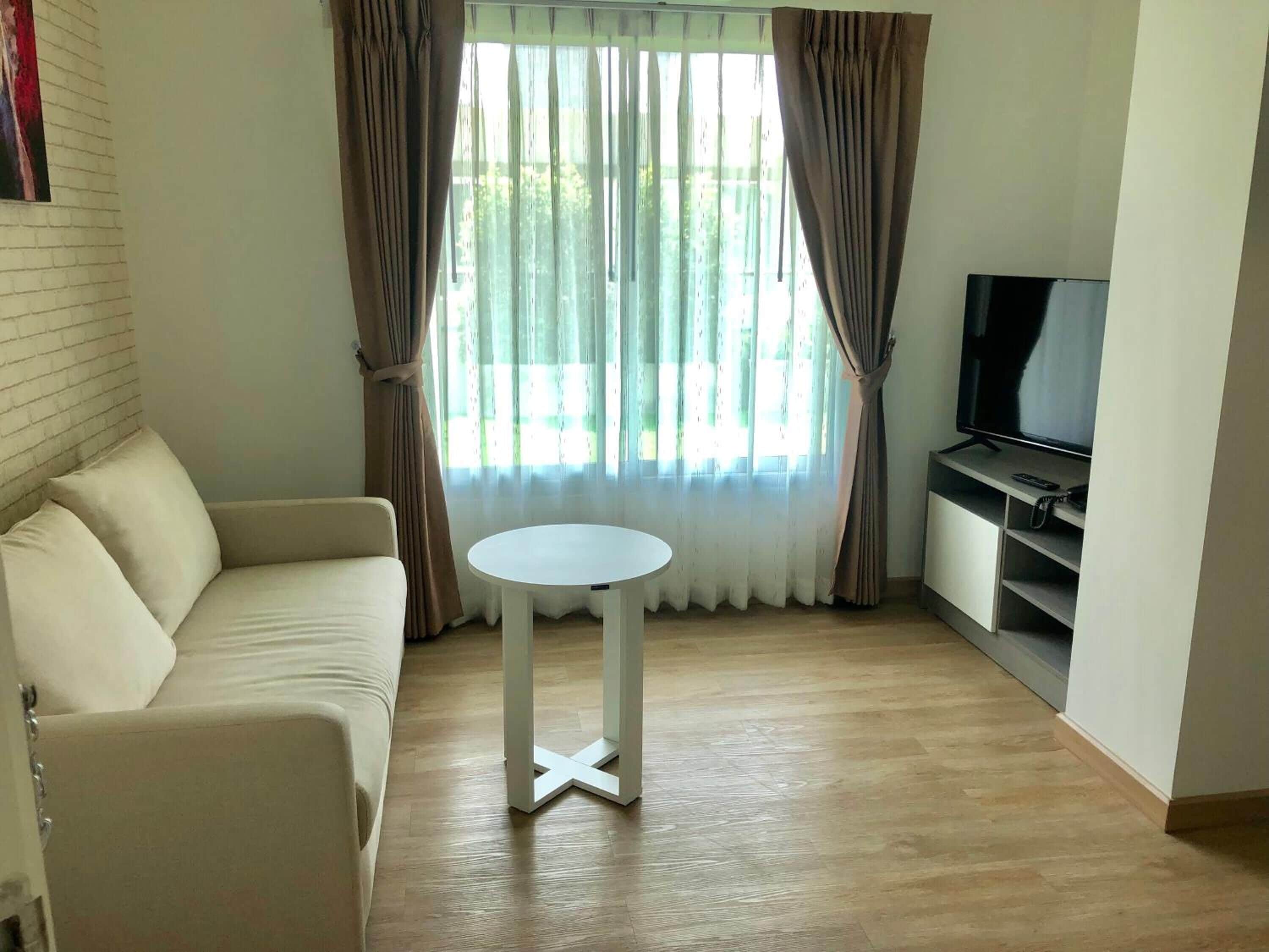 Una sala de estar con un sofá color beige, una mesita blanca, un televisor sobre un mueble bajo y una ventana con cortinas.