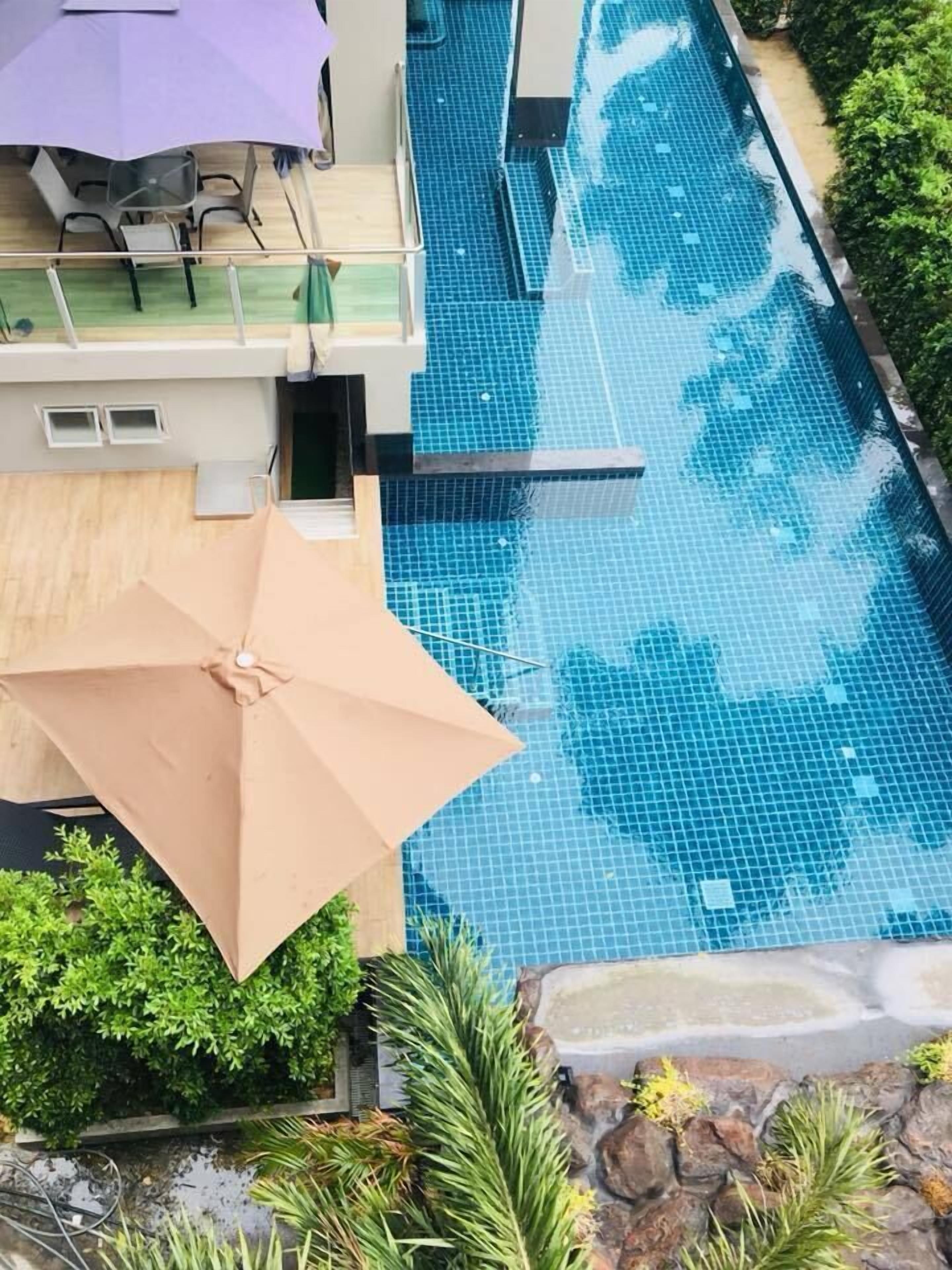 A swimming pool with a tiled deck area, a covered seating area with chairs and an umbrella, and surrounding greenery.