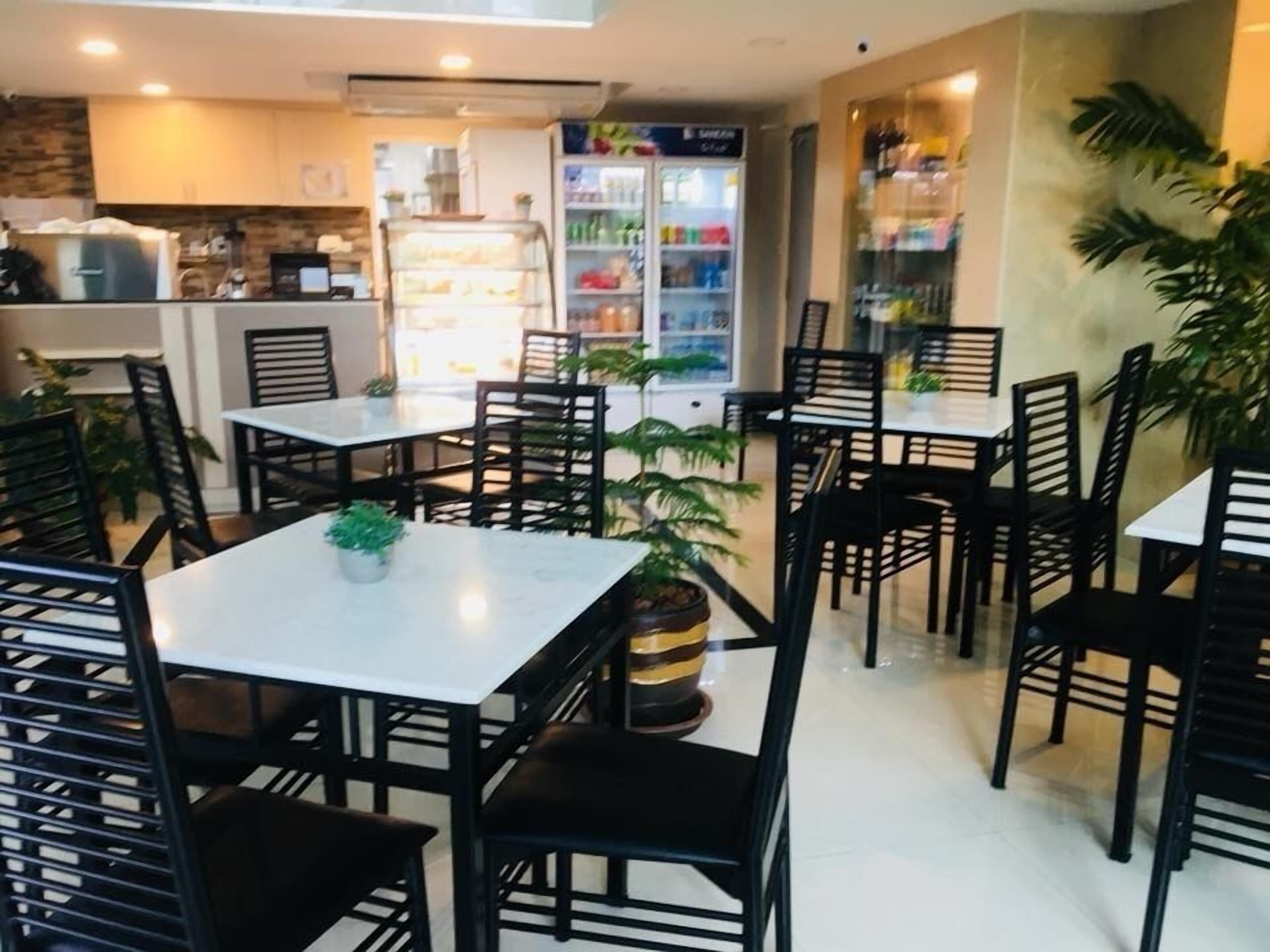 Un restaurante con sillas negras y mesas blancas, un refrigerador con bebidas y una zona de cocina al fondo.