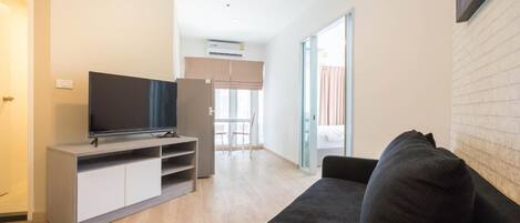 A modern living room with a flat-screen TV on a wooden stand, a dark sofa, and a sliding glass door leading to a balcony.