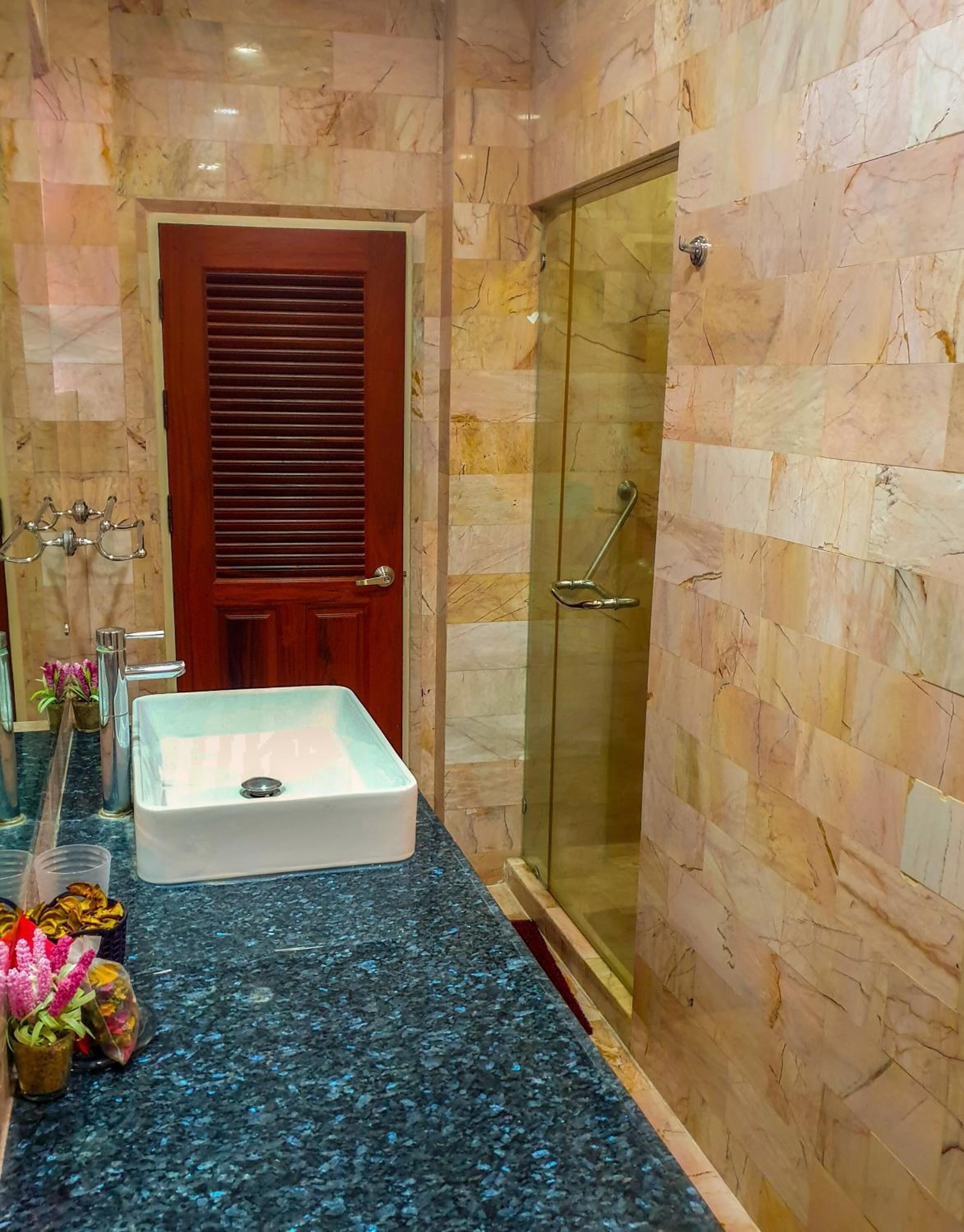 A bathroom with a marble wall, a wooden door, a glass shower enclosure, and a sink with a marble countertop.
