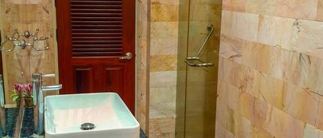 A bathroom with a marble wall, a wooden door, a glass shower enclosure, and a sink with a marble countertop.