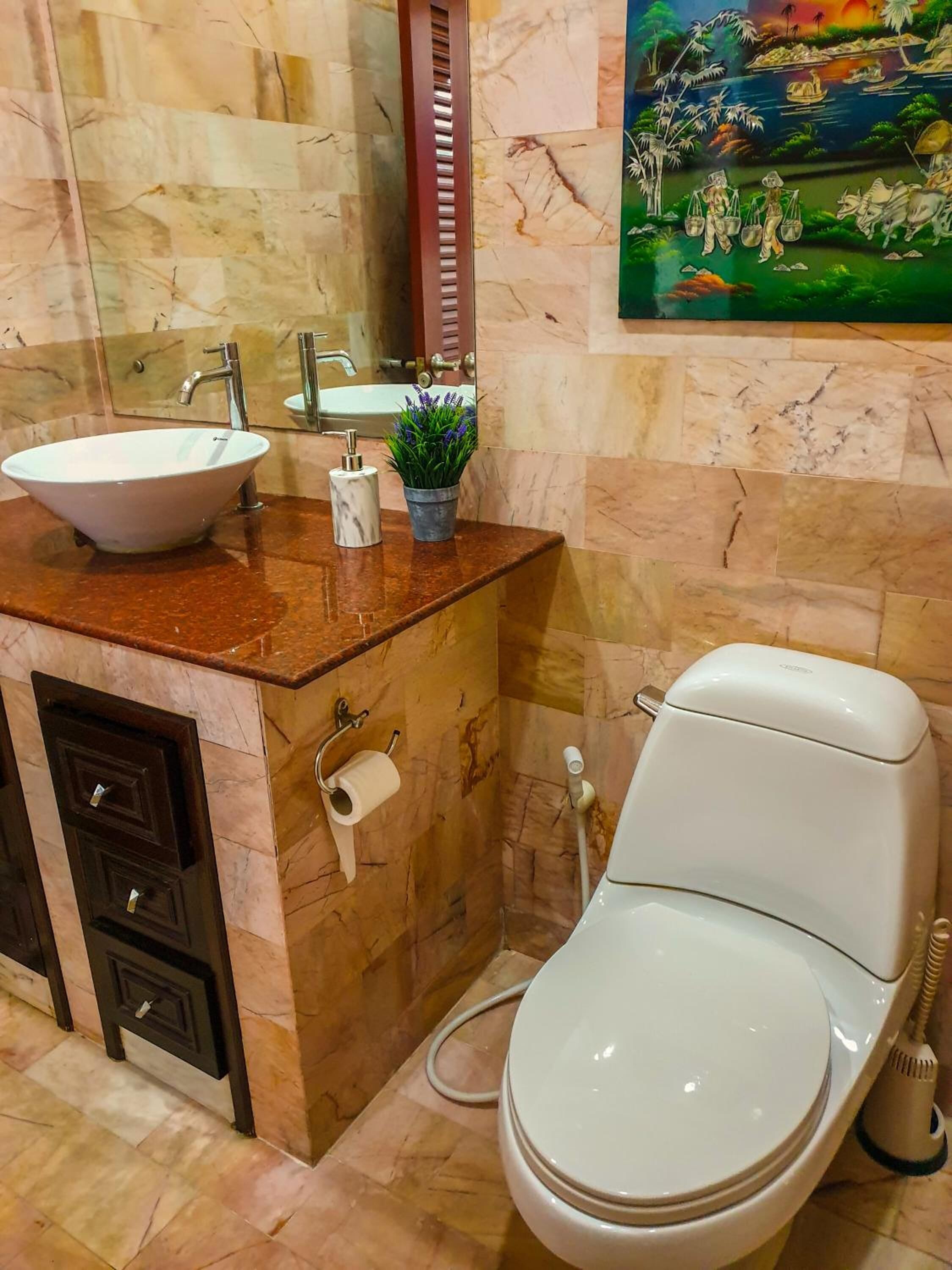 Een badkamer met een marmeren wastafel, toilet en een schilderij van een tropisch landschap.