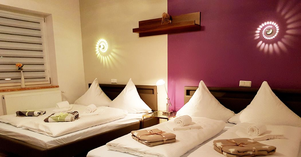 Habitación de hotel con dos camas, cada una con ropa de cama y almohadas blancas, una mesita de noche con lámpara y un estante de pared con un objeto decorativo.