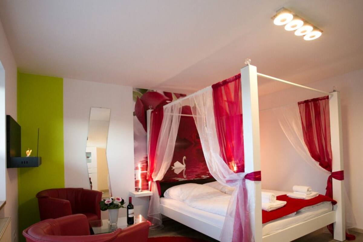 A four-poster bed with red and white drapes, a bedside table with a bottle and flowers, and a red armchair.