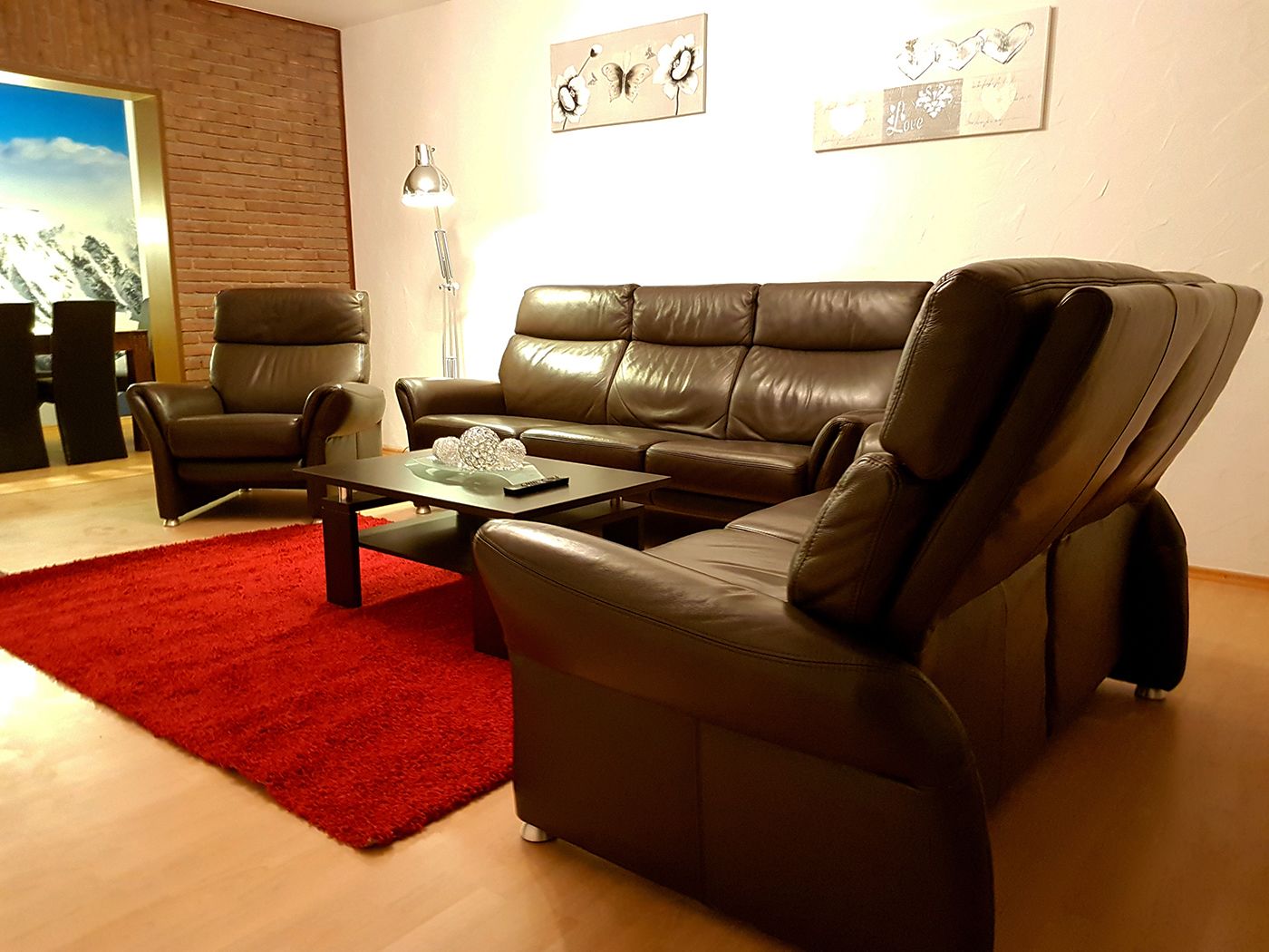 A modern living room with a brown leather sofa, two armchairs, a coffee table, and a red rug. There are wall art pieces and a lamp.