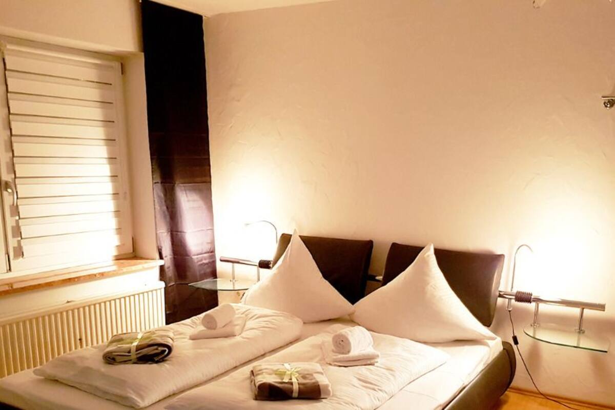 A neatly made bed with pillows and towels, a bedside table with a lamp, and a window with blinds.