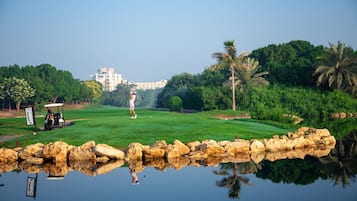 Un campo de golf con un estanque al frente, palmeras y un edificio en el fondo.