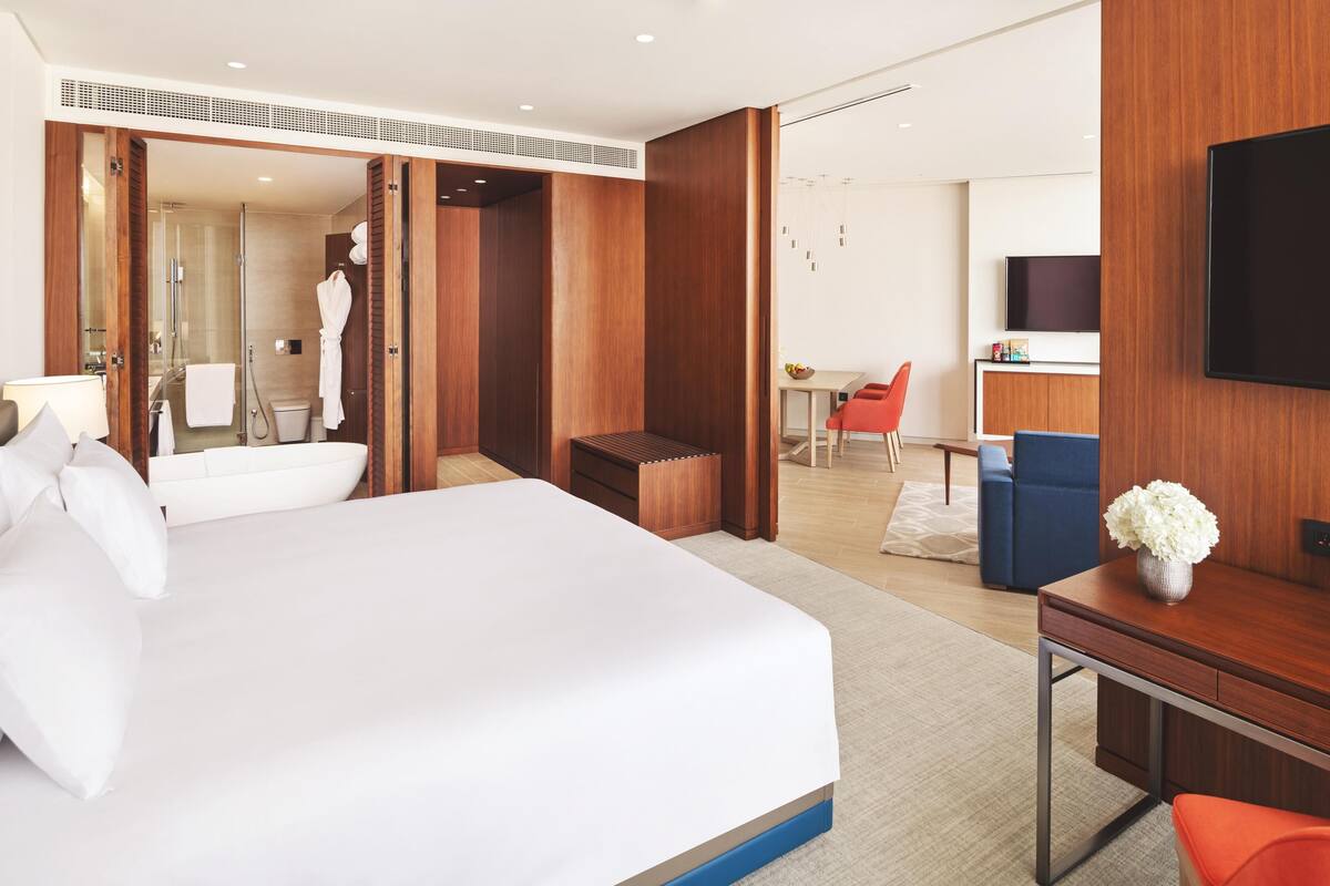 A modern hotel room with a large bed, a desk, a TV, a shower, and a dining area.