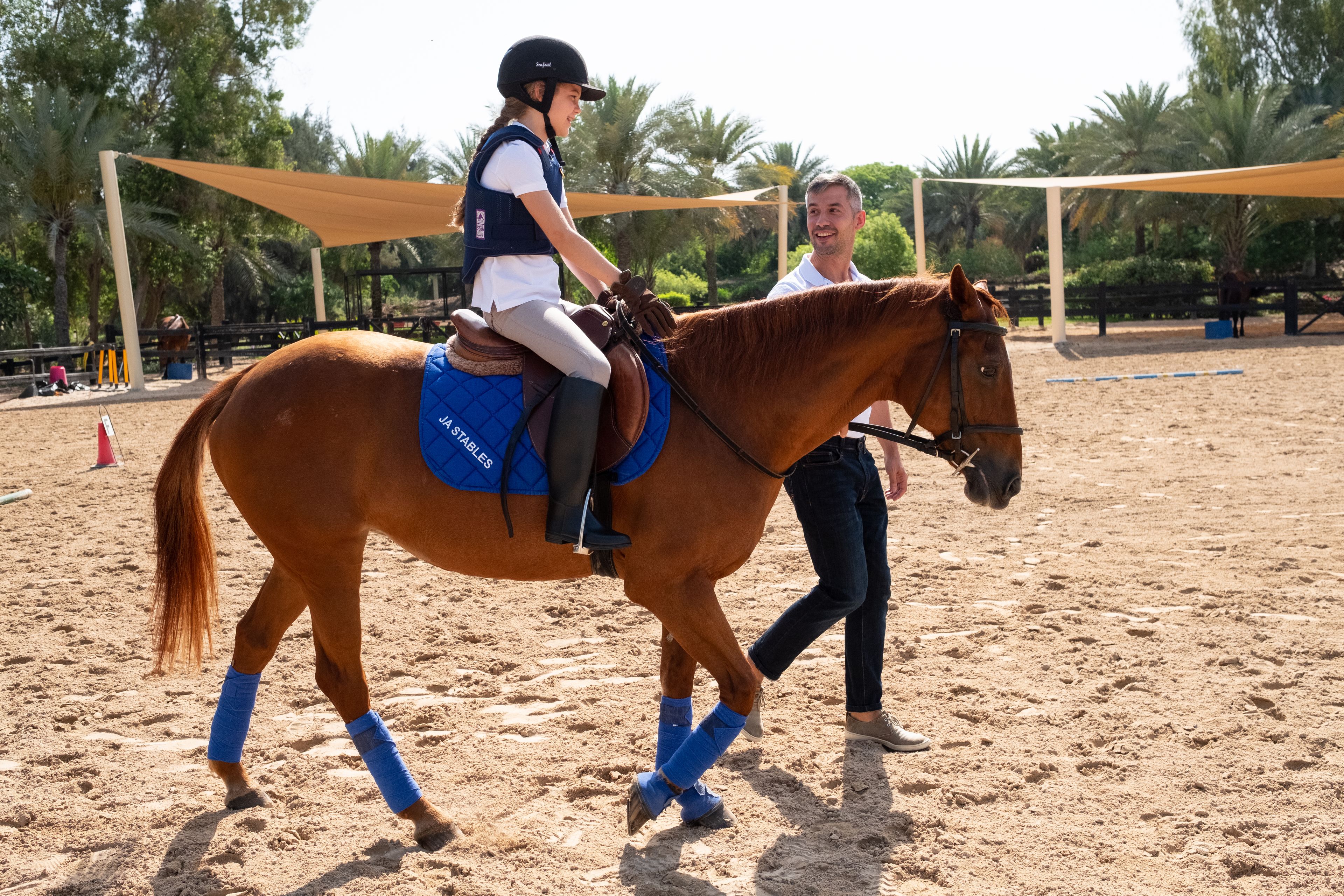 A person riding a horse in an outdoor arena with a sandy ground, surrounded by trees and a shaded area.