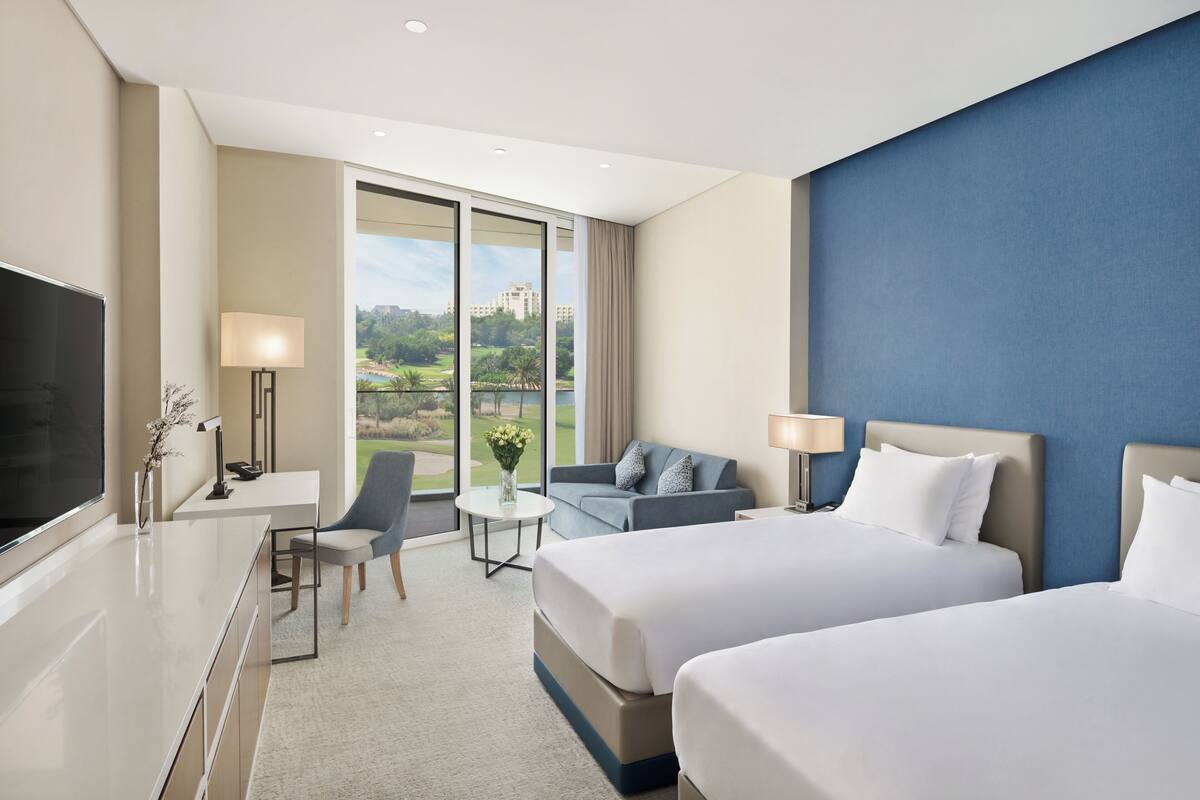 A modern hotel room with two beds, a desk, a chair, a TV, and a view of a golf course.