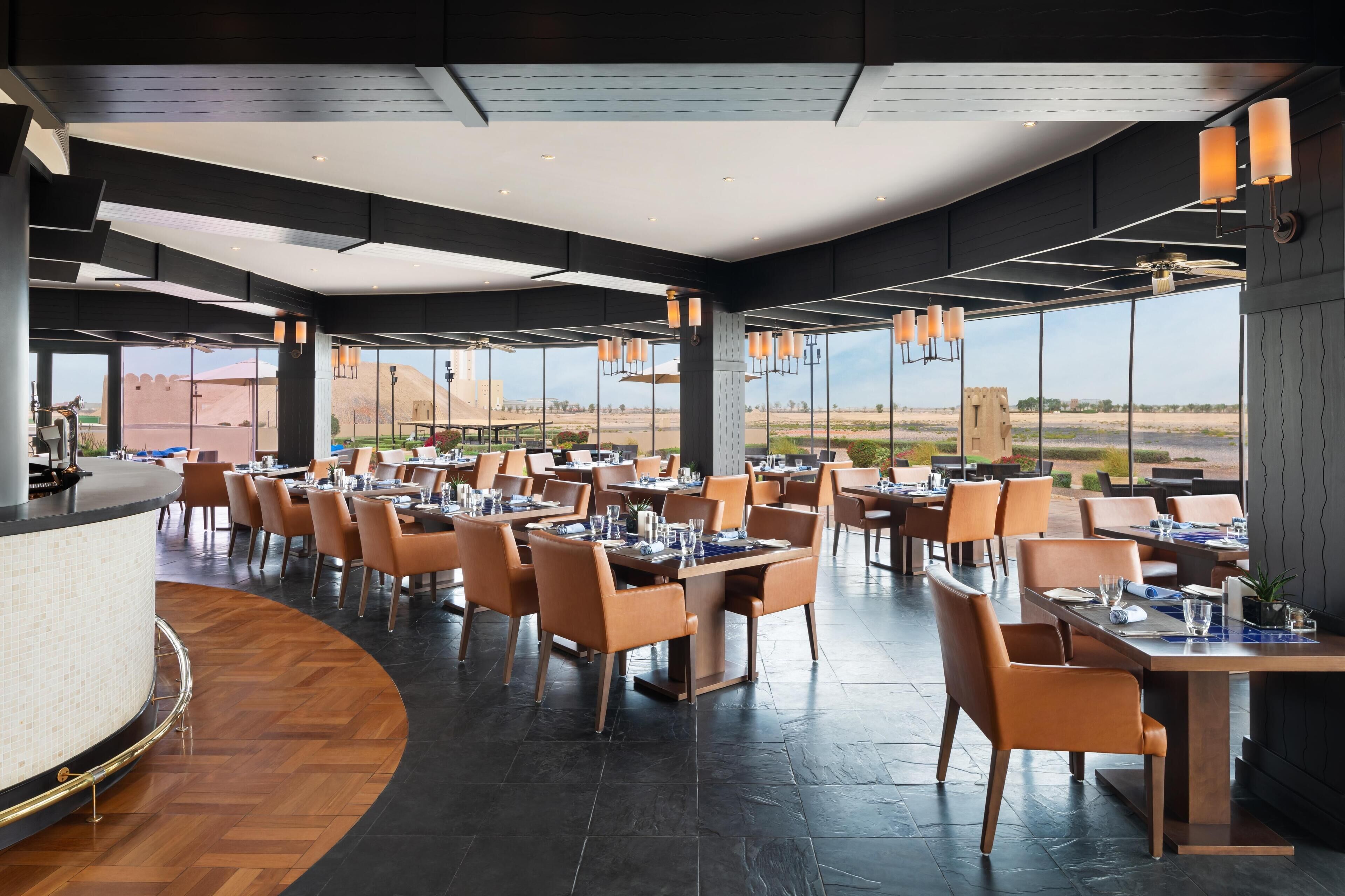 Ein modernes Restaurant mit einer gewölbten Theke, mehreren Esstischen mit Stühlen und großen Fenstern mit Blick auf die Wüstenlandschaft.