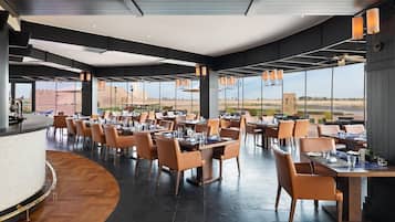 Ein modernes Restaurant mit einer gewölbten Theke, mehreren Esstischen mit Stühlen und großen Fenstern mit Blick auf die Wüstenlandschaft.
