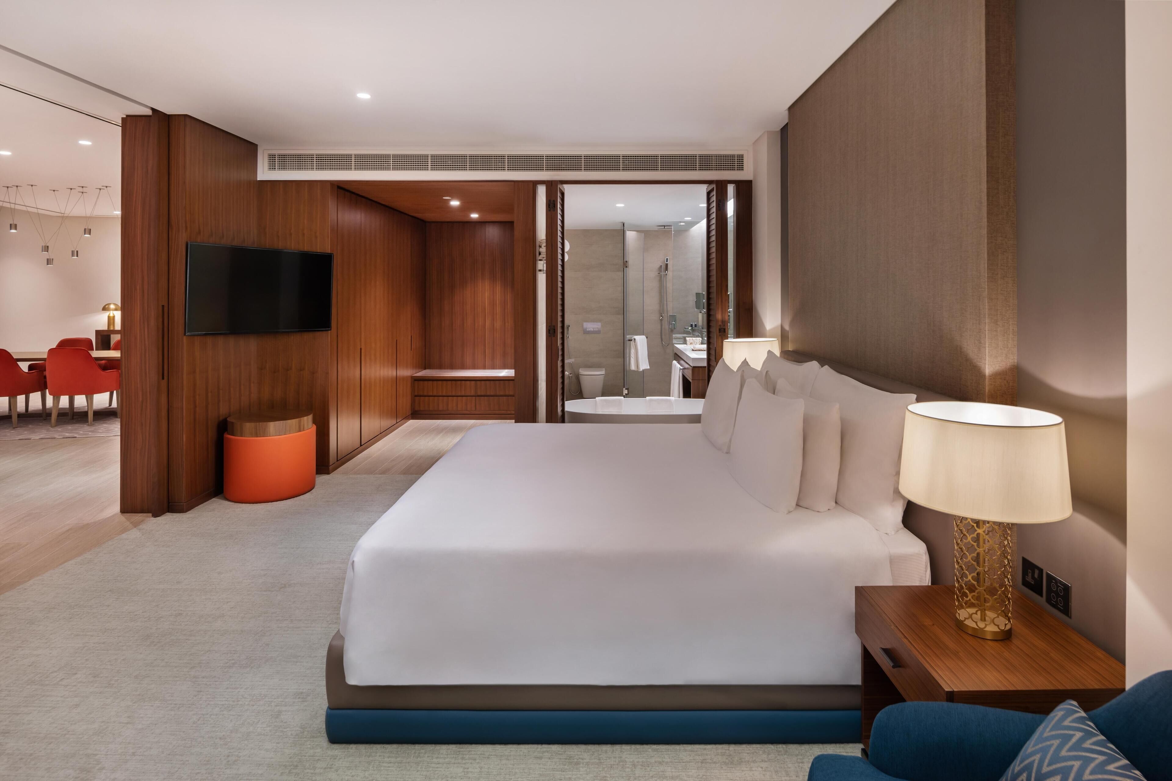 A modern hotel room with a large bed, a TV, a desk, and a bathroom visible through an open door.