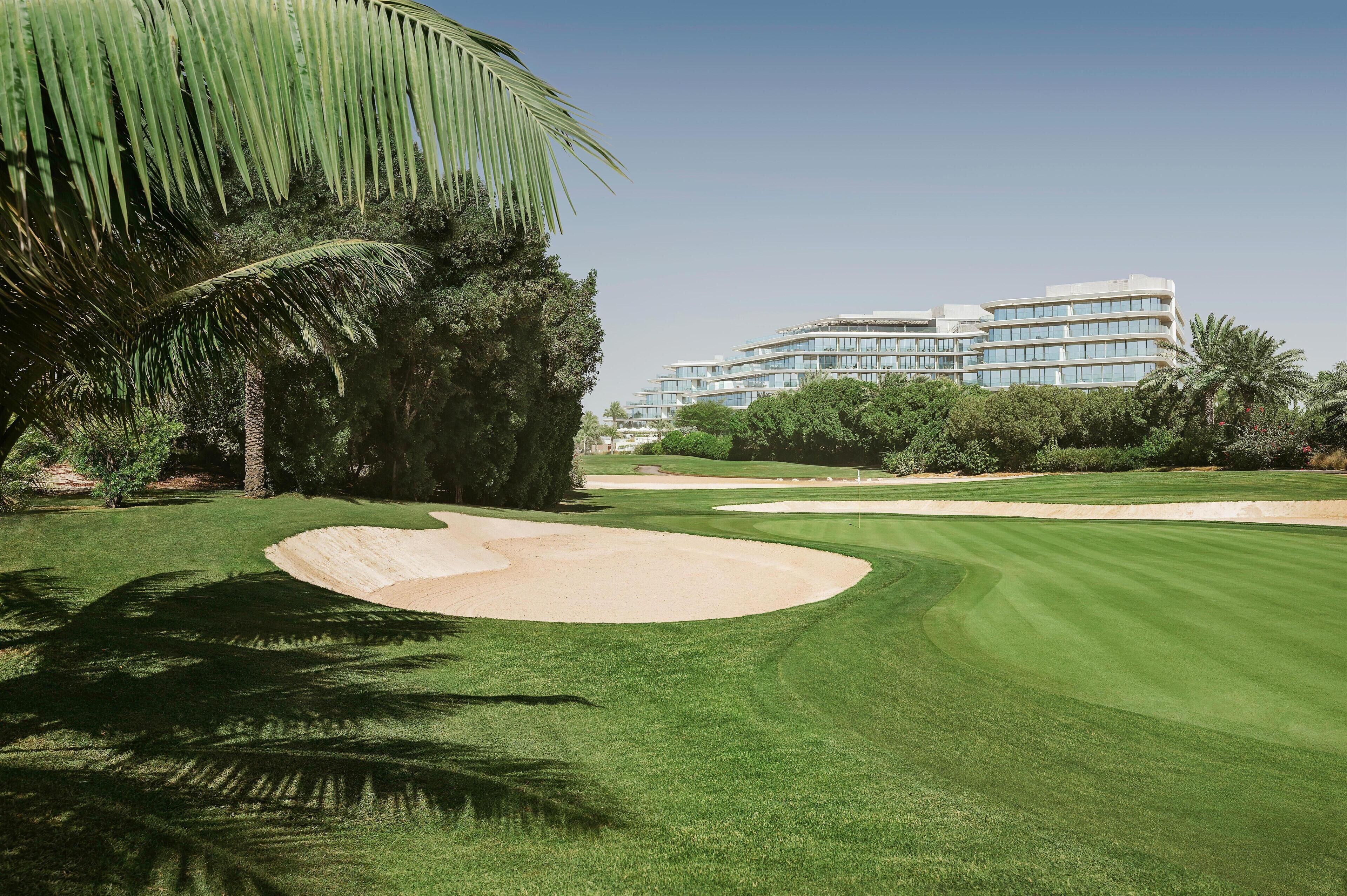 A golf course with a sand bunker, palm trees, and a modern building in the background.