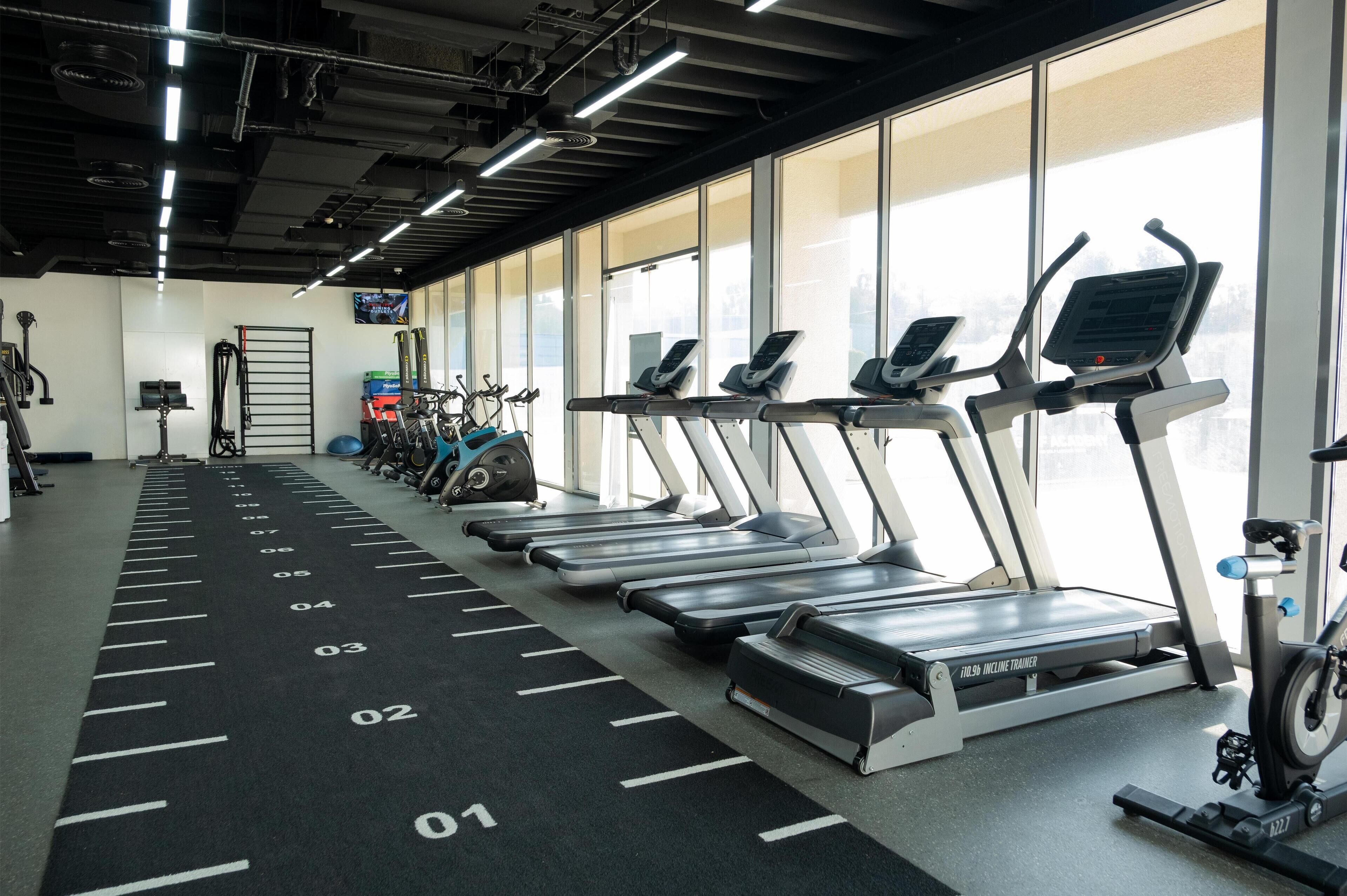 Une salle de sport avec des tapis de course alignés en rangées, un vélo d’appartement et des ballons d’exercice.