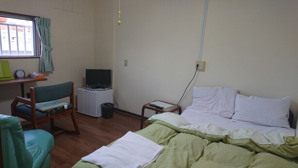 Une chambre d’hôtel avec un lit, une chaise, un petit bureau avec une télévision et une fenêtre avec des rideaux.