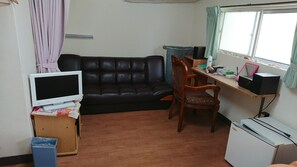 Un salon comprenant un canapé en cuir noir, une chaise en bois, un bureau avec un ordinateur, un téléviseur et un réfrigérateur.