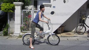 Une personne à vélo circule dans une rue, avec un bâtiment blanc et une clôture en métal en arrière-plan, le tout agrémenté de fleurs.