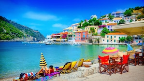 A coastal scene with colorful buildings, a beach with lounge chairs and umbrellas, and boats in the water. - Villa Artemis (Kefalonia)