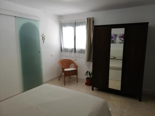 Una habitación con cama, una silla, planta en maceta, ventana con cortinas y puerta corredera.